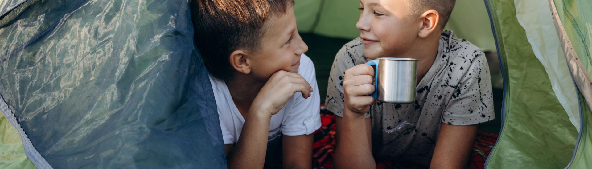 Two kids in a tent, one drinking from a cup.